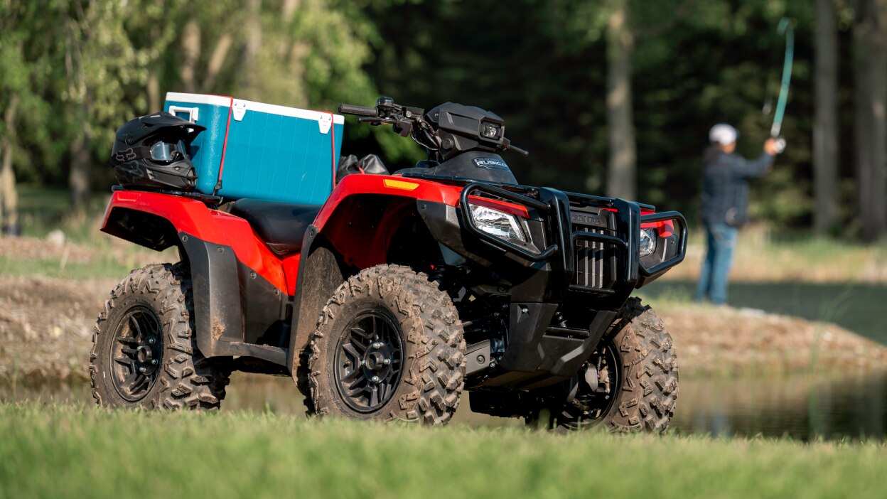 A Honda Rubicon 700 parked with a blue cooler strapped to the back with a man fishing in the background. 