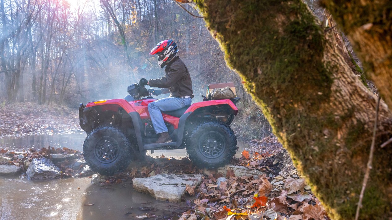A rider about to go into a large puddle in a wooded area on a Honda Rubicon 700. 