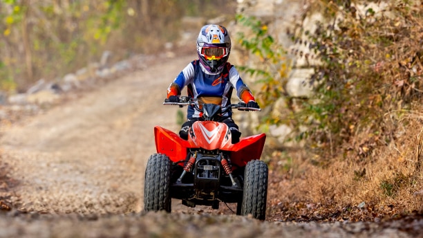 Young rider on ATV on dirt road. Grass and fence in background.