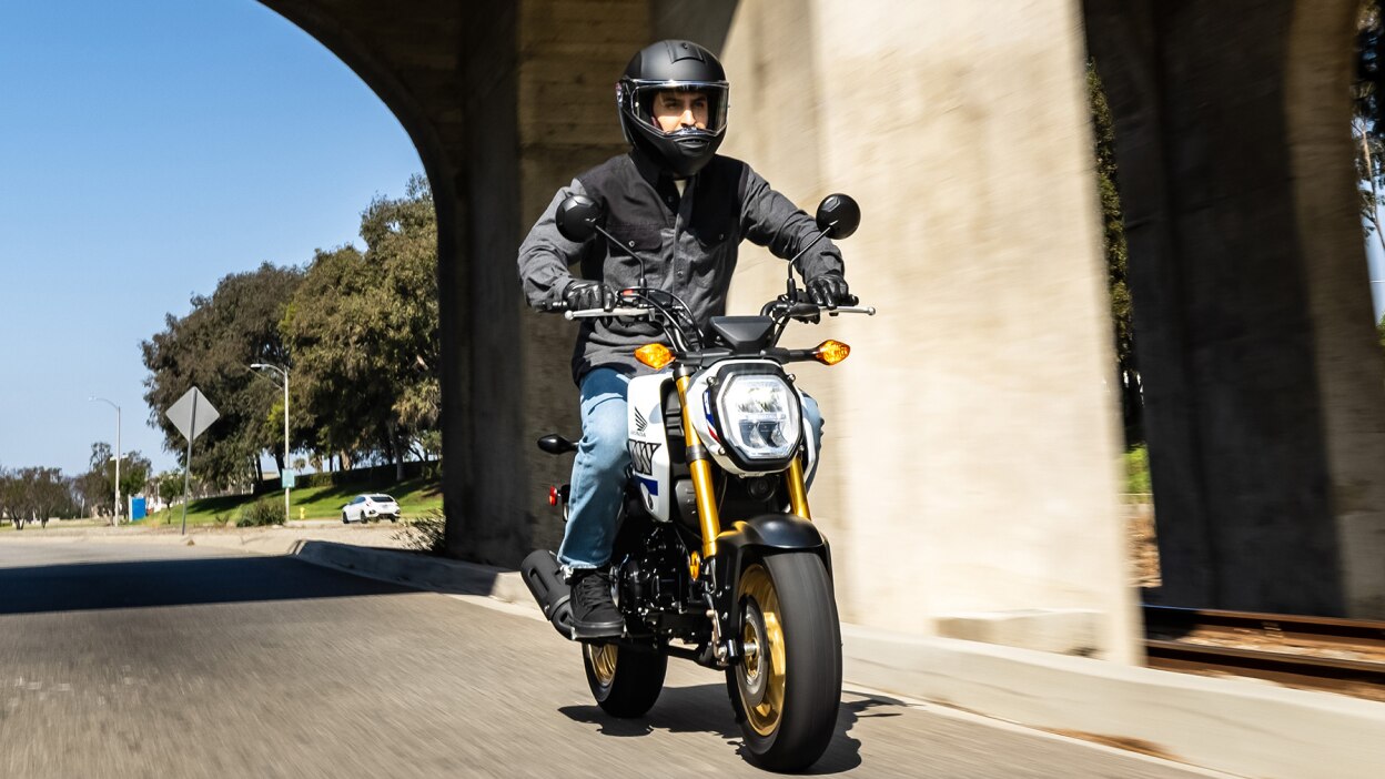 Rider on yellow bike at slight right angle driving on road. Bridge above and trees and white car in background.