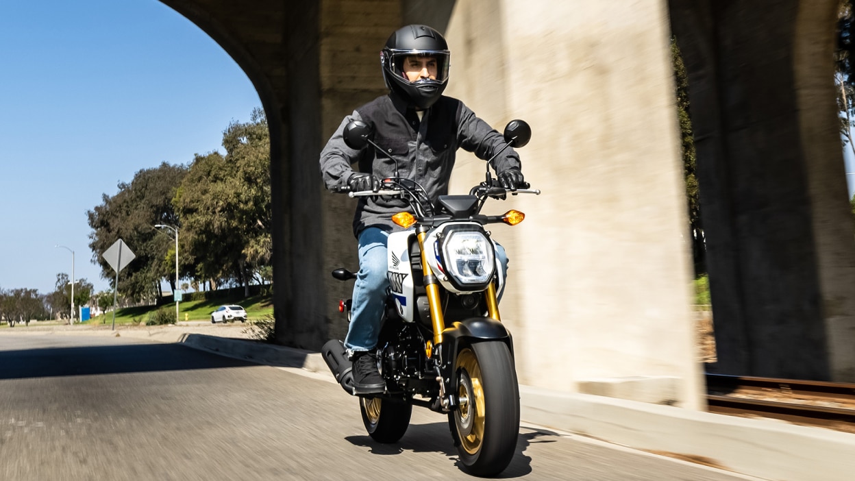 Rider on yellow bike at slight right angle driving on road. Bridge above and trees and white car in background.