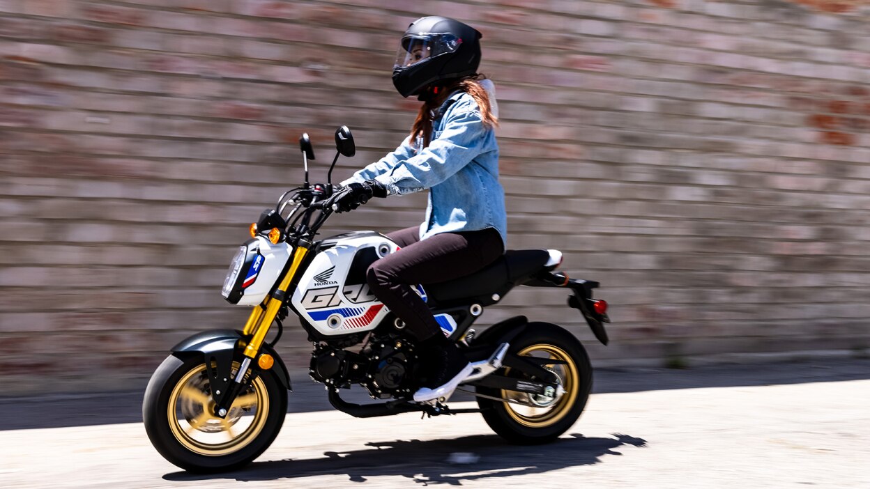 A rider with light blue denim jacket going left on street. Bricked wall in background.