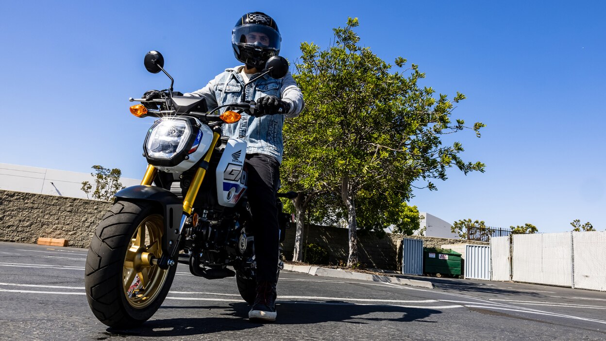 Rider with light blue denim jacket parked on bike in car park. Trees, walls and clear sky in background.