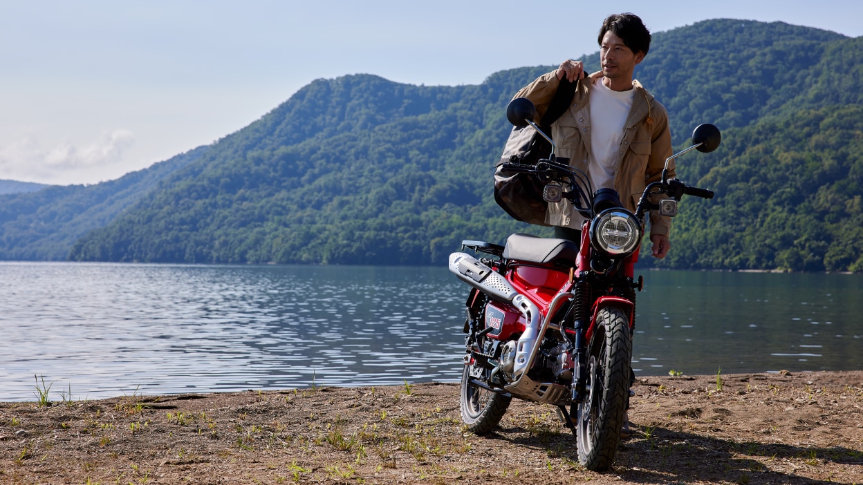 Rider with no helmet standing to the left of red bike. Lake and forest landscape in background.