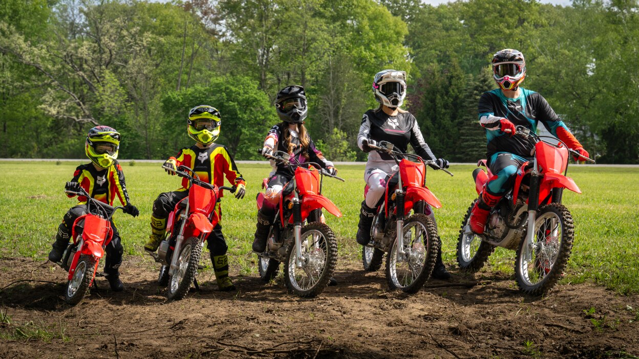 Two adult riders, three young riders stationary side by side. Greenary in background.