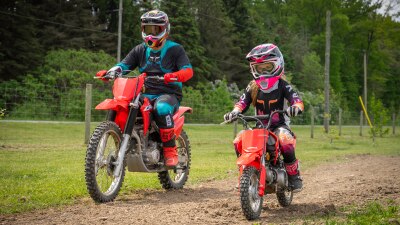 Adult rider and  young rider going right on dirt track. Green field, trees and fence in background.