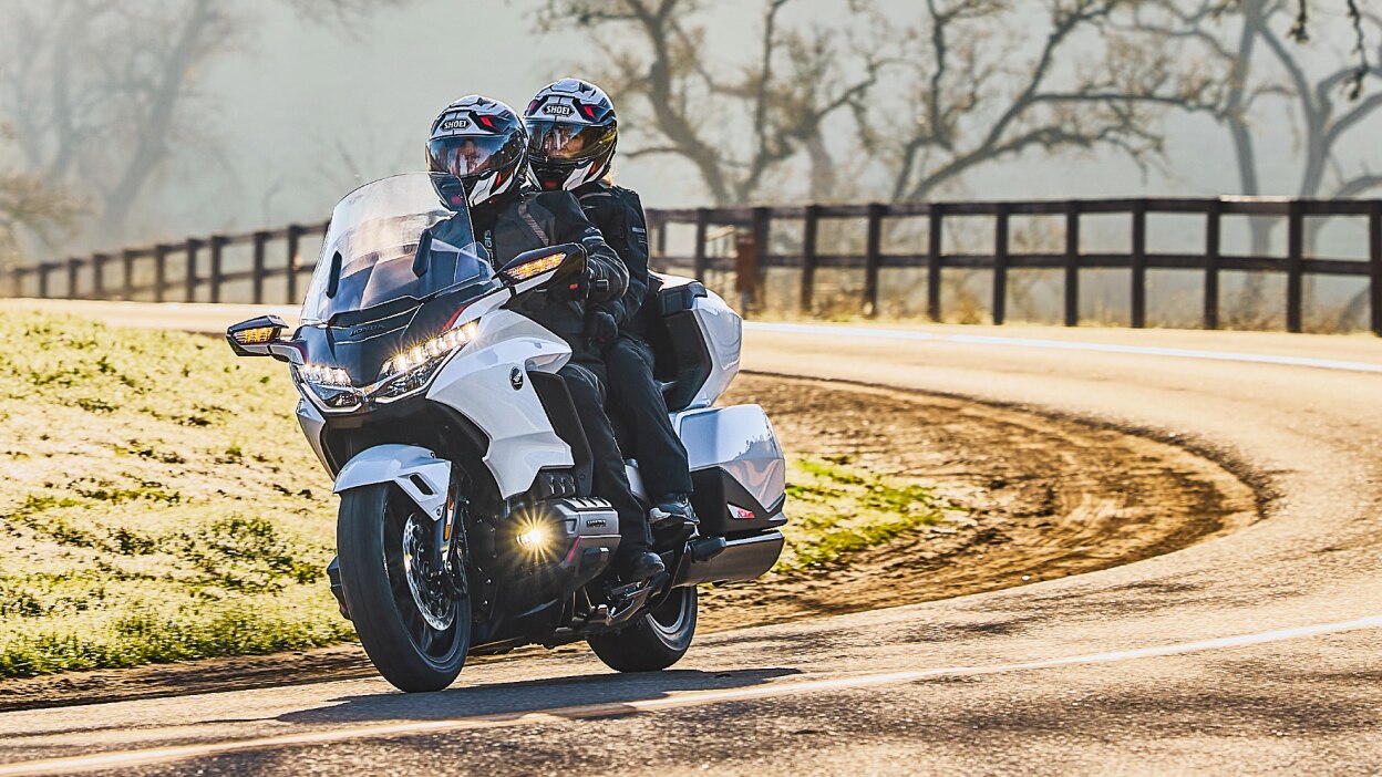 Two riders on white bike going left. Trees and fence in background.