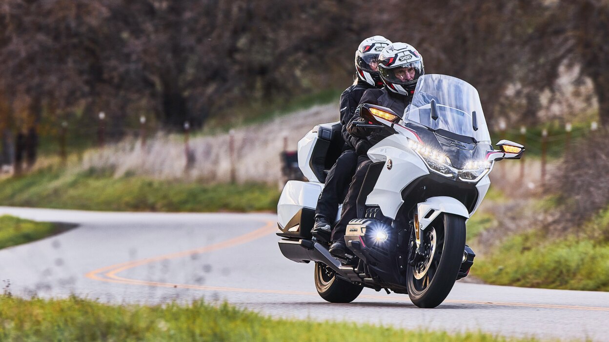 Two riders on white bike going right. Trees in background.