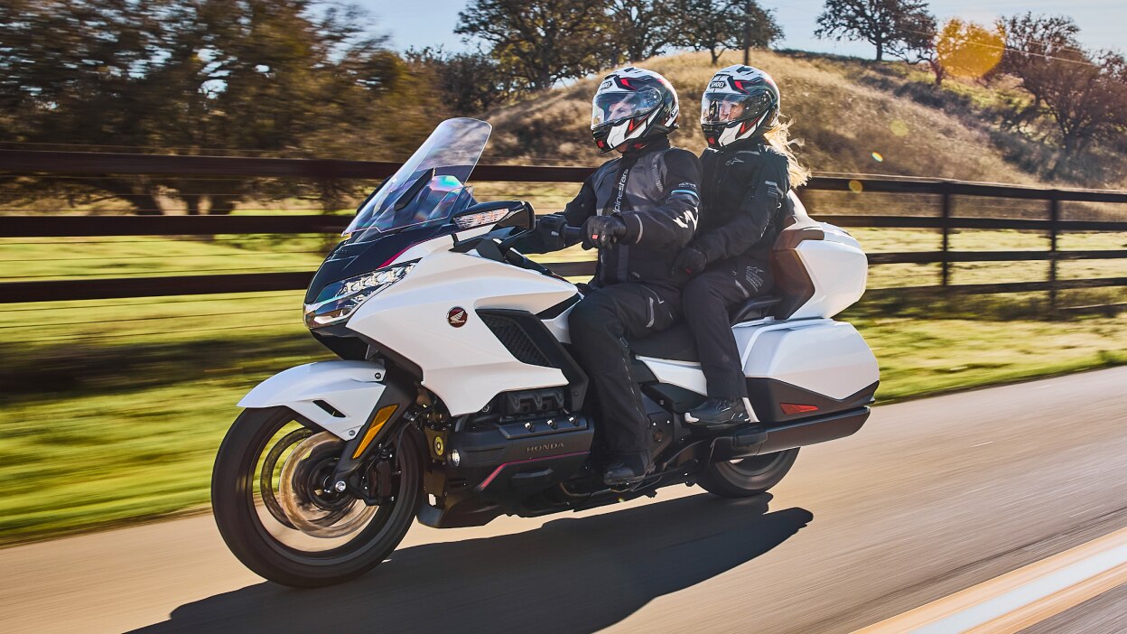 Two riders on white bike going left on road. Bushes, fence and trees in background.