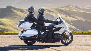 Two riders on white bike going right. Green Hills in background.