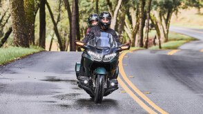 Two riders on green bike on road. Front facing. Tress in the background on the left.