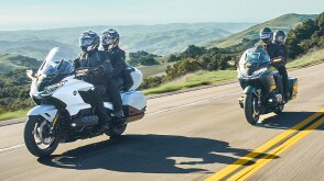 Two riders on white bike infront of two riders on green bike on road. Hills in background.