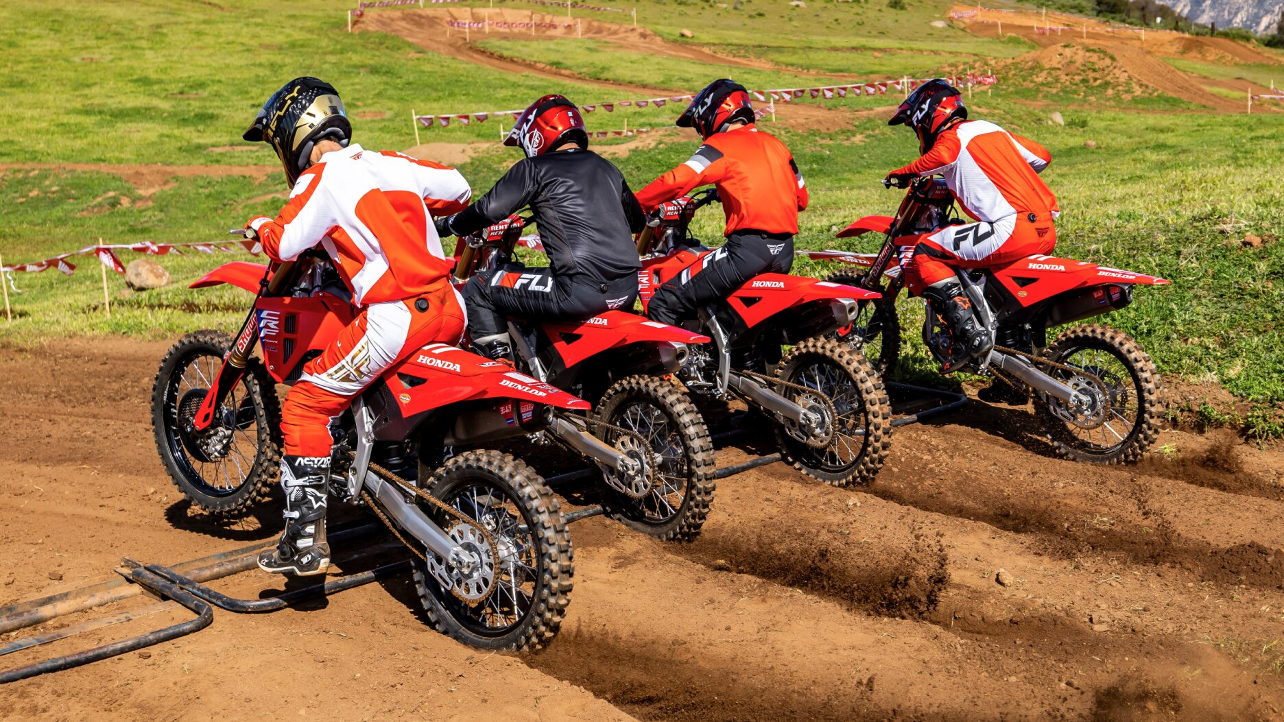 Four riders lines up about to start on dirt track. Back angle.