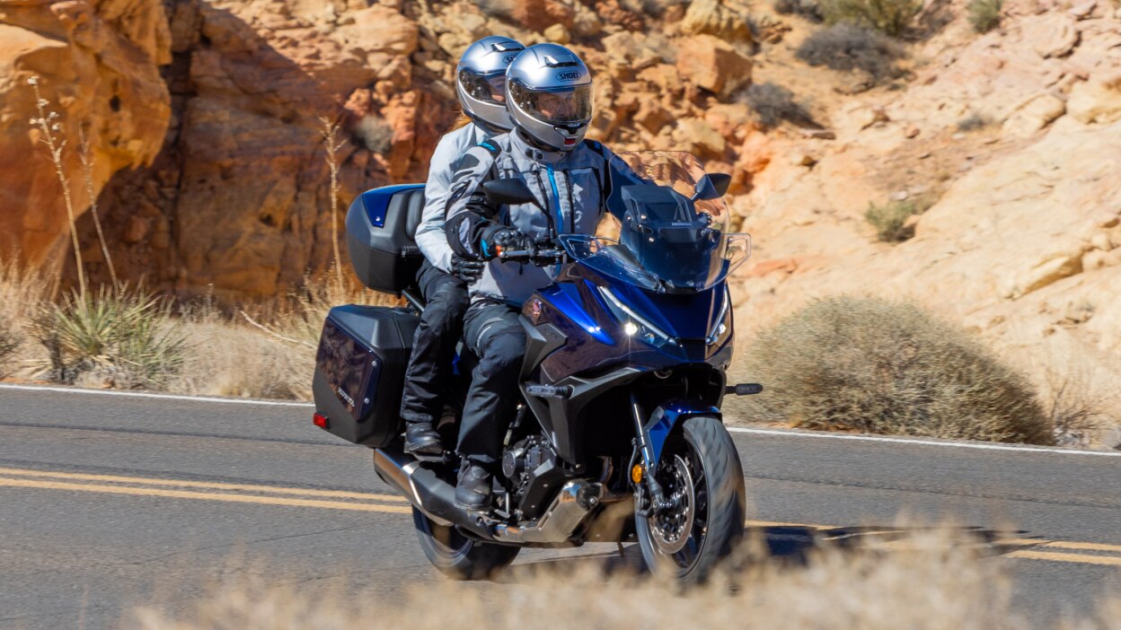 Rider and passenger facing right on NT1100. Desert landscape in background.