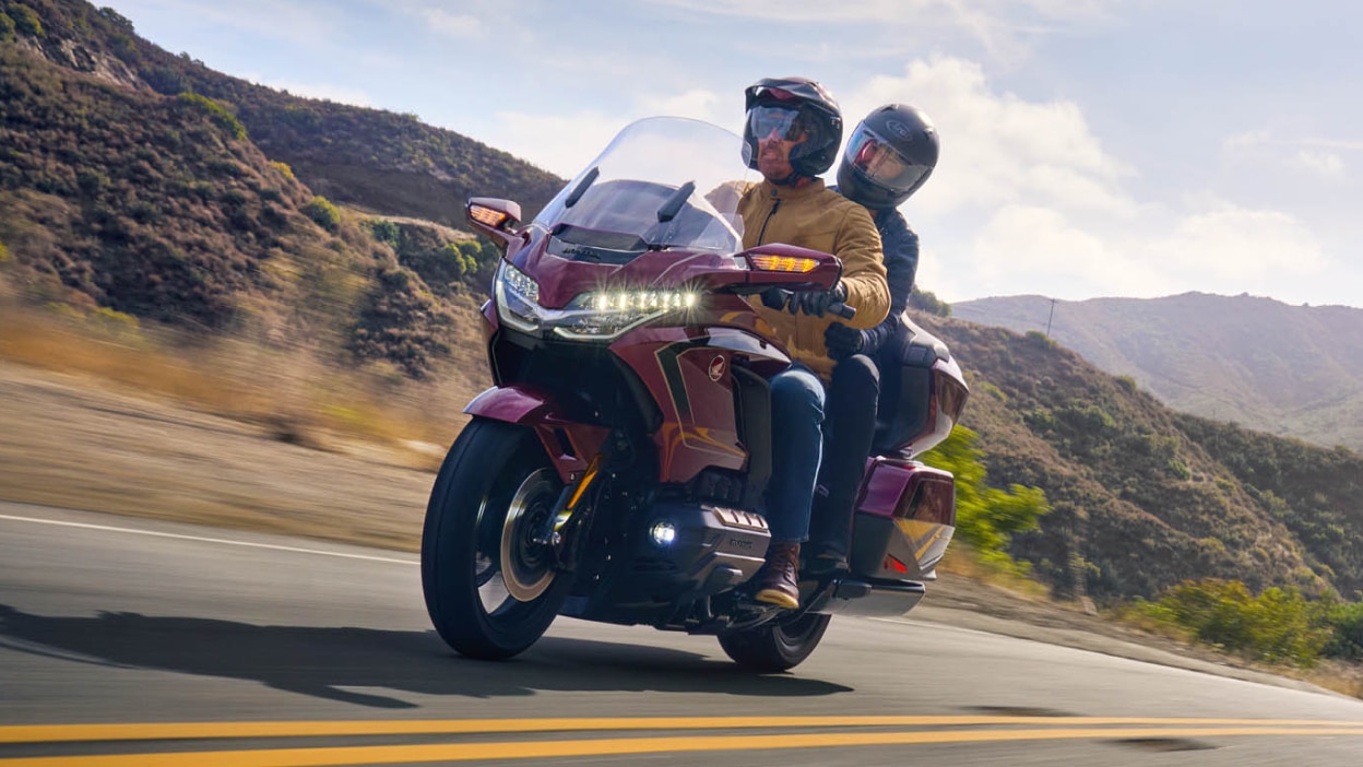 Rider and passenger on red Gold Wing Tour going left on road. Mountains in background.