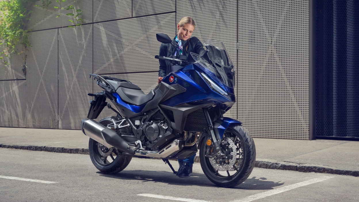 NT1100 is stationary on the road. Grey building in the background. A woman behind bike.