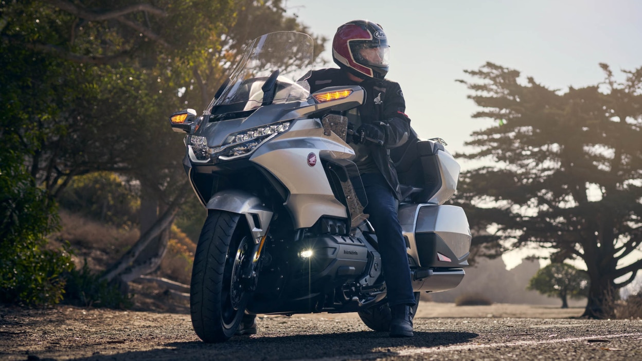 Rider stationary on silver Gold Wing Tour facing right. Trees in background.
