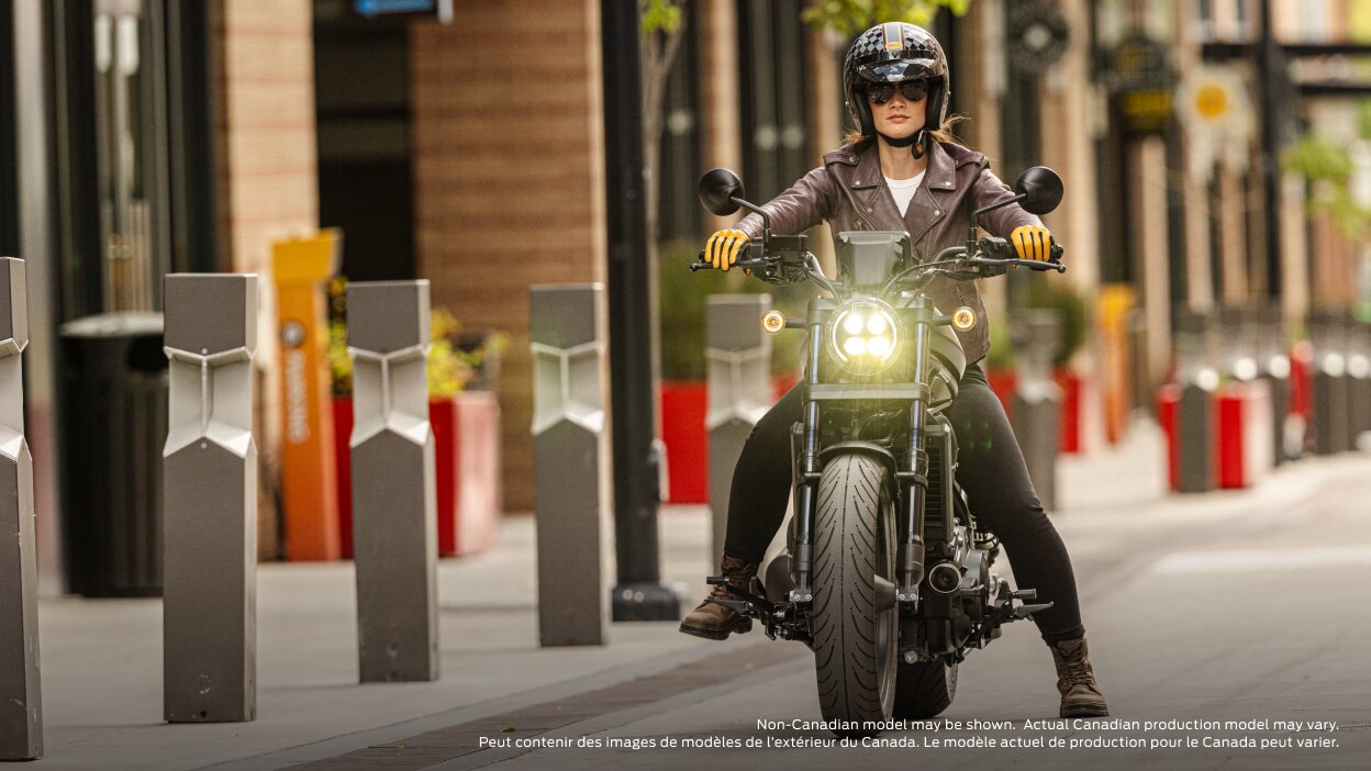 Front facing of rider on bike on road with headlight on. Side wall with pillars to the left. Non-Canadian model(s) may be shown.  Actual Canadian production model(s) may vary.