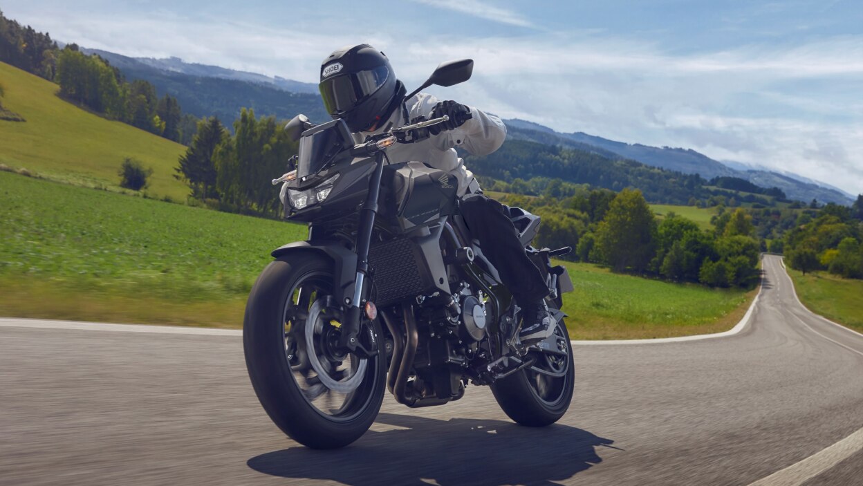 Rider on CB750, turning right on road, front facing. Green hills, trees and grass in background
