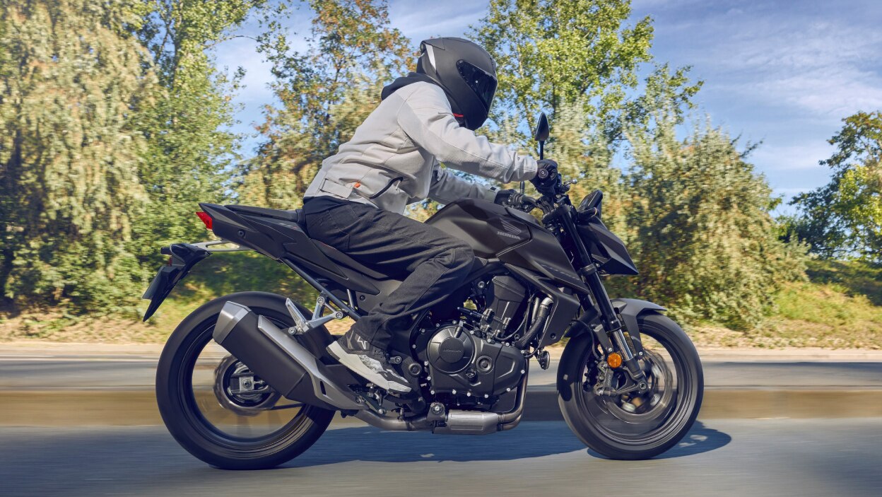 Rider on CB500F going right. Green trees and bushes in background.