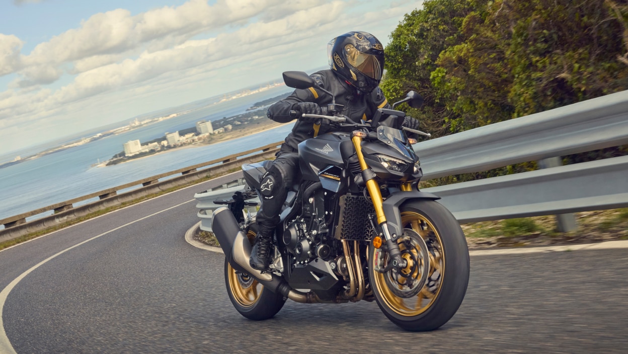 Rider making left turn on CB1000SP. Sea and city in background.