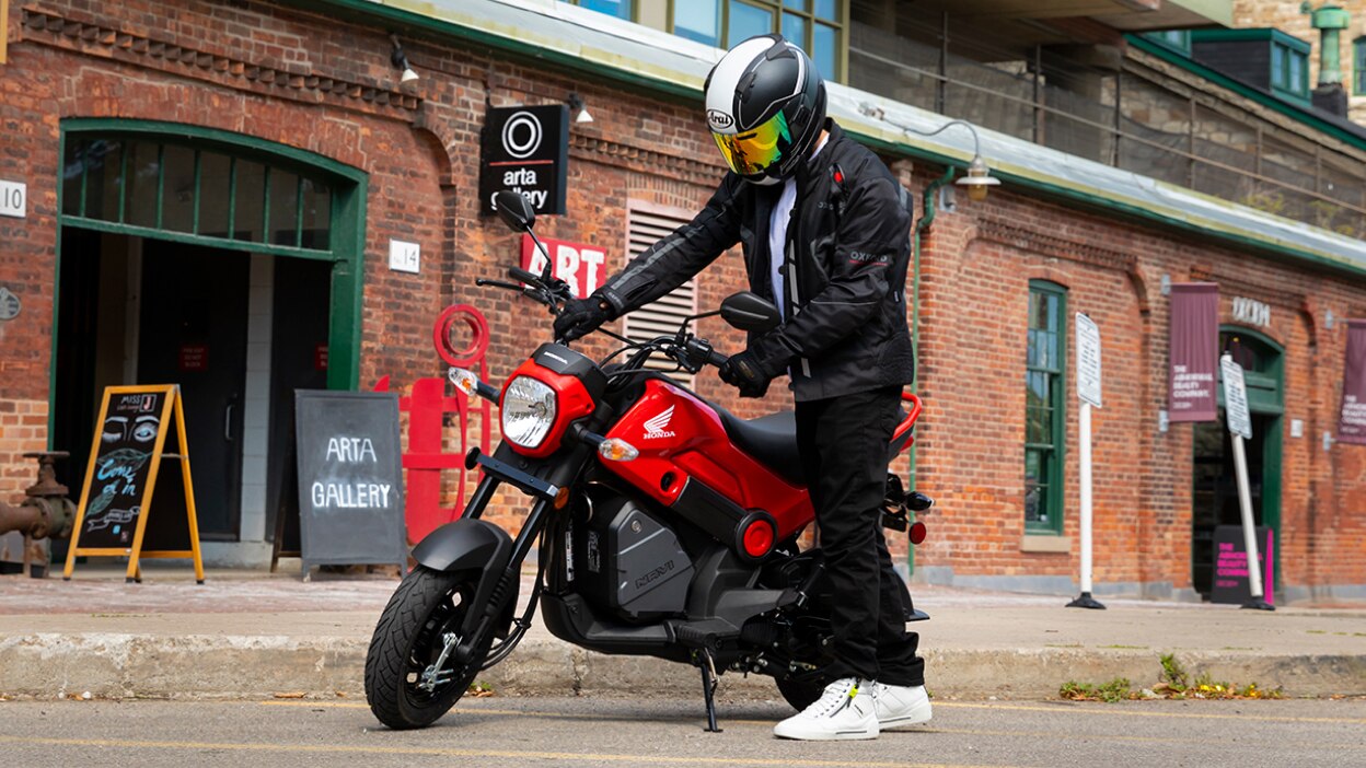 Person on a red Honda Navi motorcycle in front of a brick building with flowers