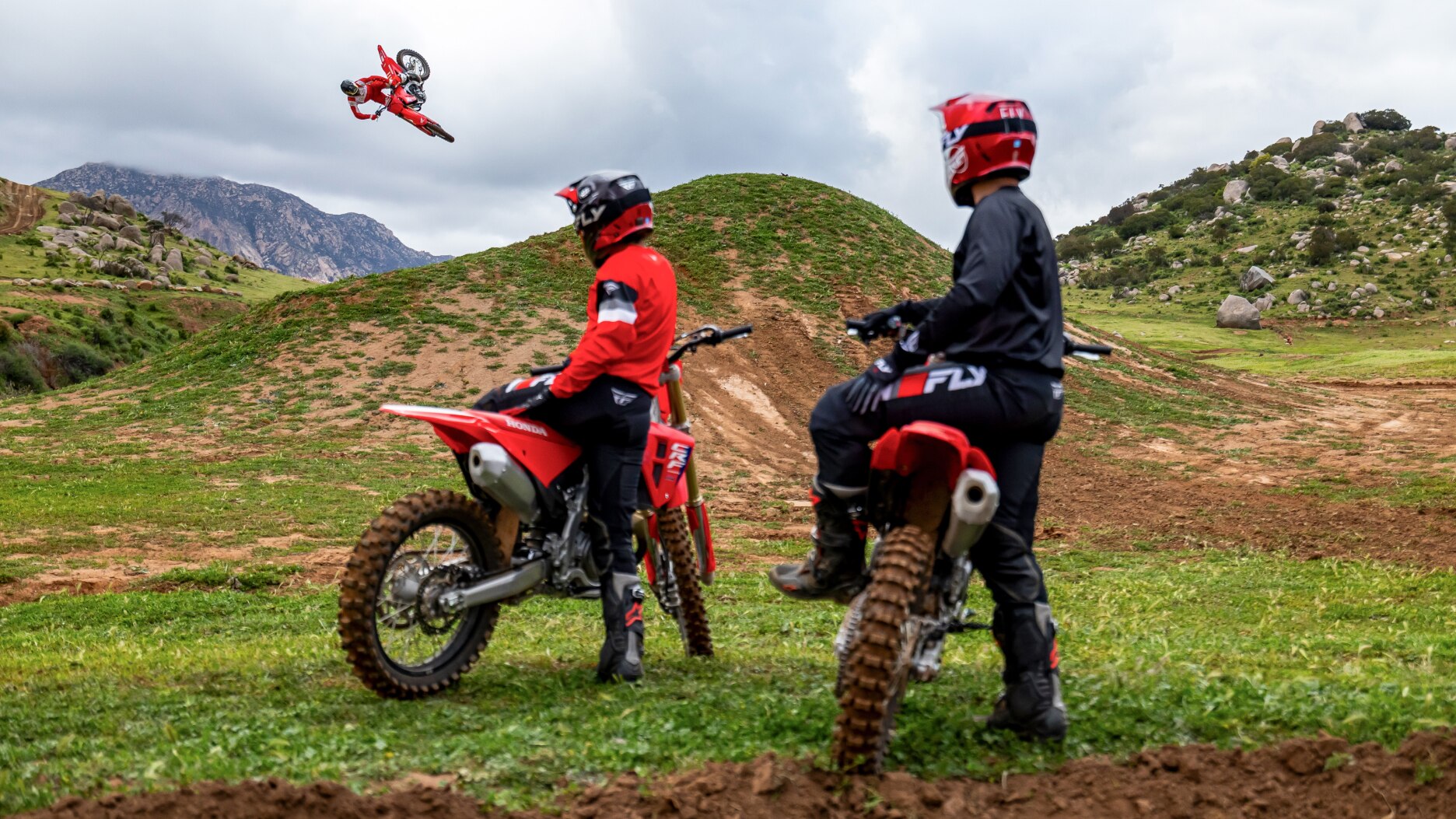 Rear view of two riders sitting on bikes looking at another rider mid air.