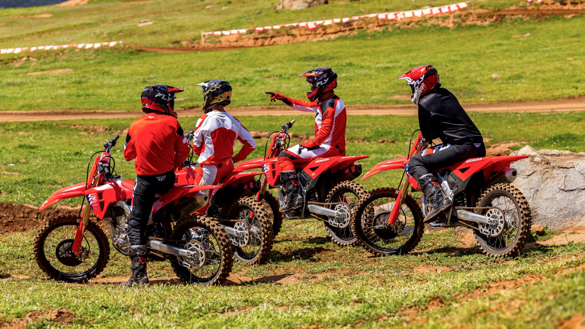 Four riders sitting on motorbikes facing left on dirt track.