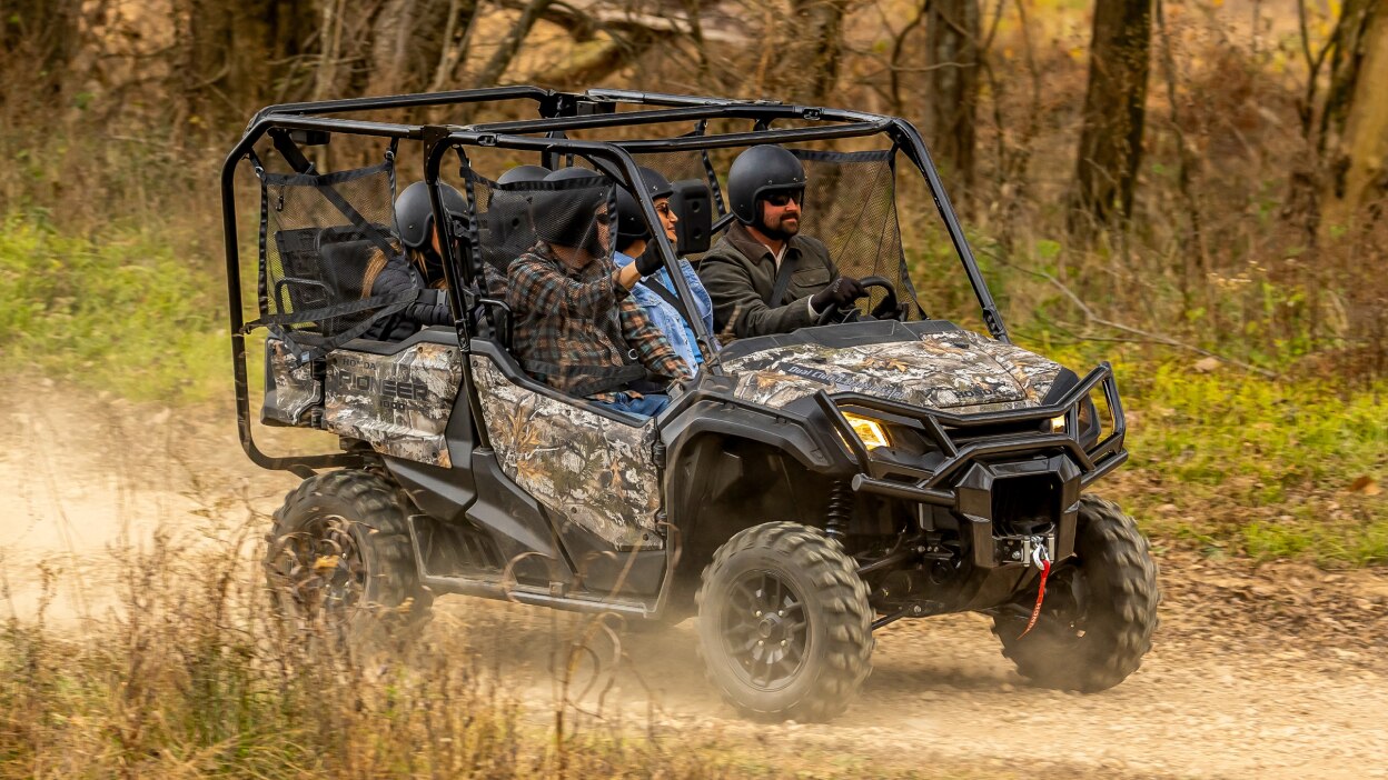 Four people in camo sxs going right. Trees in background.