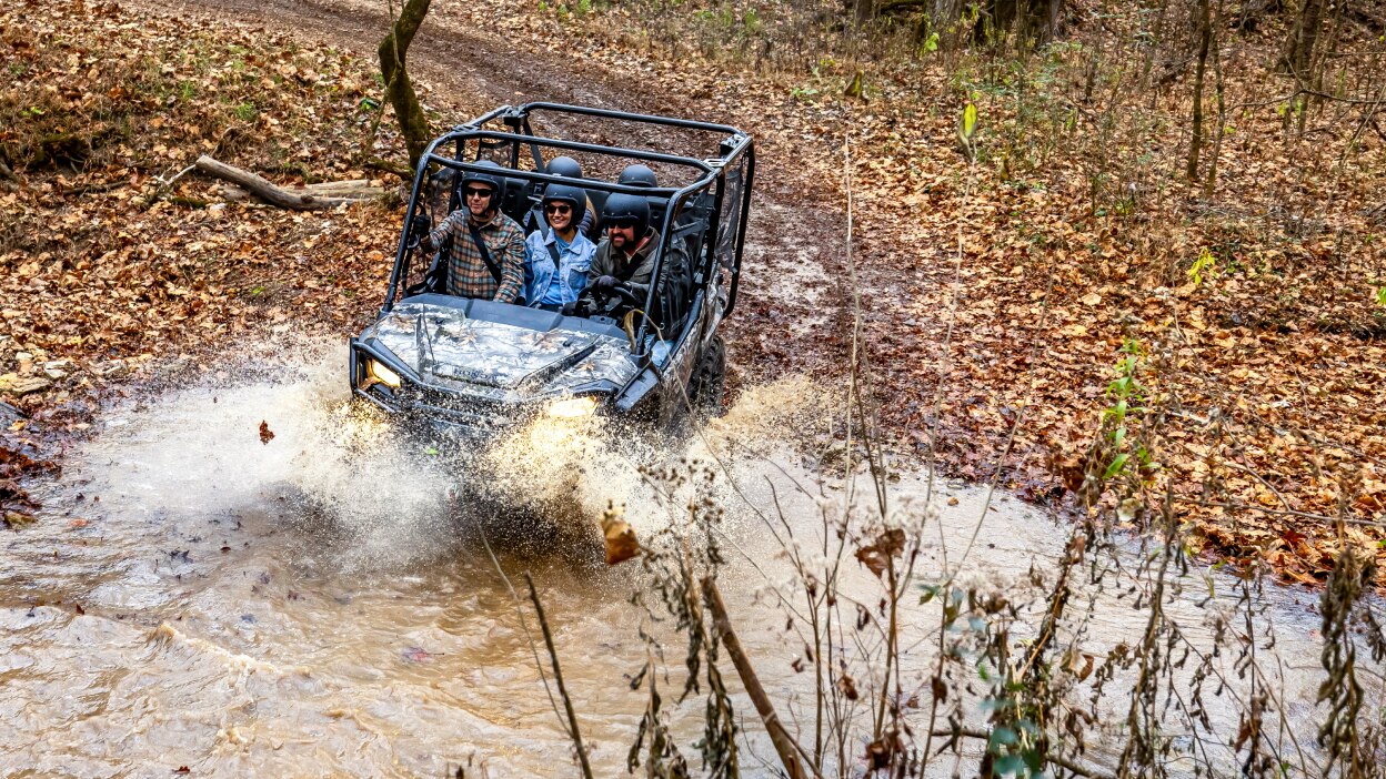 Four people in camo sxs going down in to muddy water. Leafy forrest background.