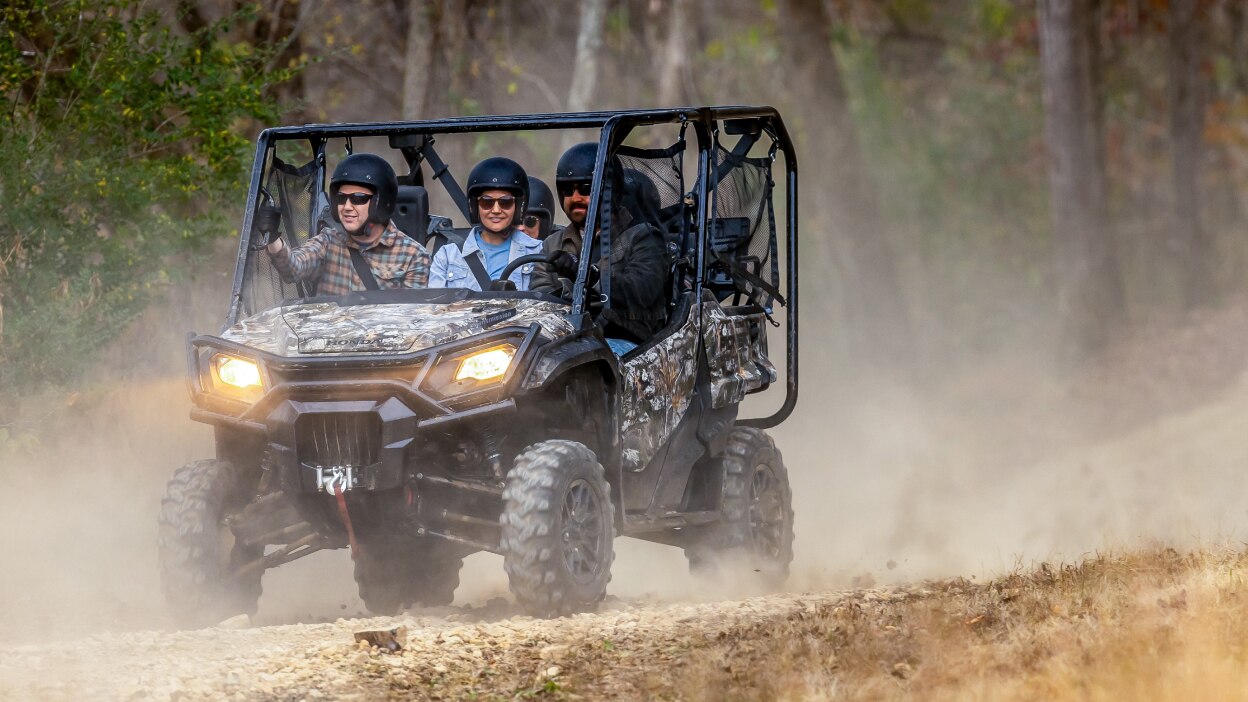 Four people in camo sxs going left on dirt track. Forrest background.