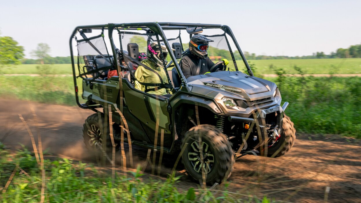 Two people in grey sxs going left. Green bushes in background.