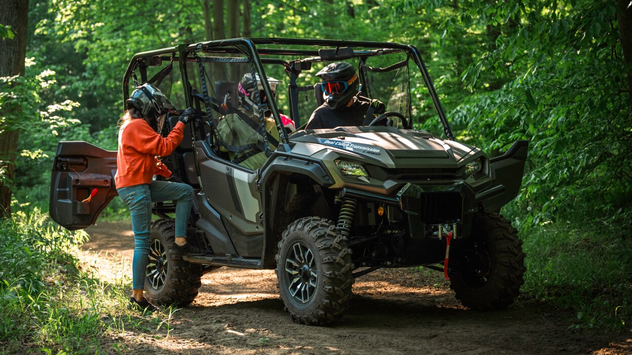 Passenger in orange jacket getting into sxs. Green forrest background.